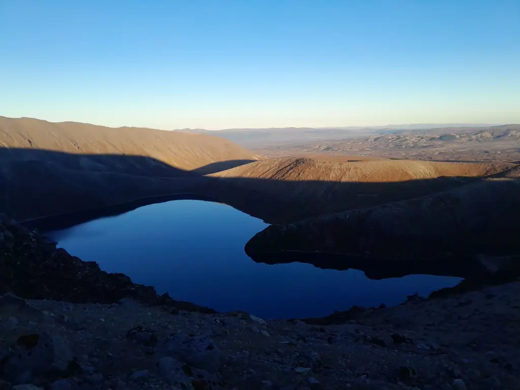 View across the serene, deep blue water of Upper Tama Lake from its surrounding, barren volcanic shoreline under a clear sky in Tongariro National Park.