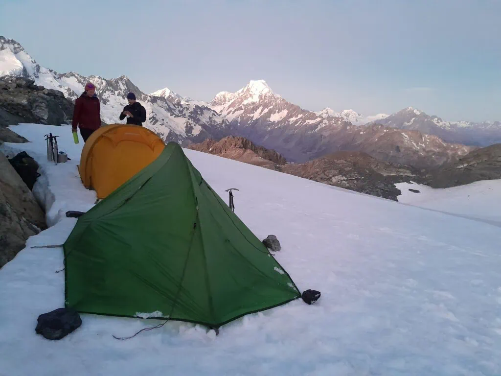 A green and yellow tent pitched on the snow of the Annette Plateau with two hikers behind them and ice-axes dug into the snow. Aoraki/Mt Cook visible in the background, Aoraki/Mt Cook National Park
