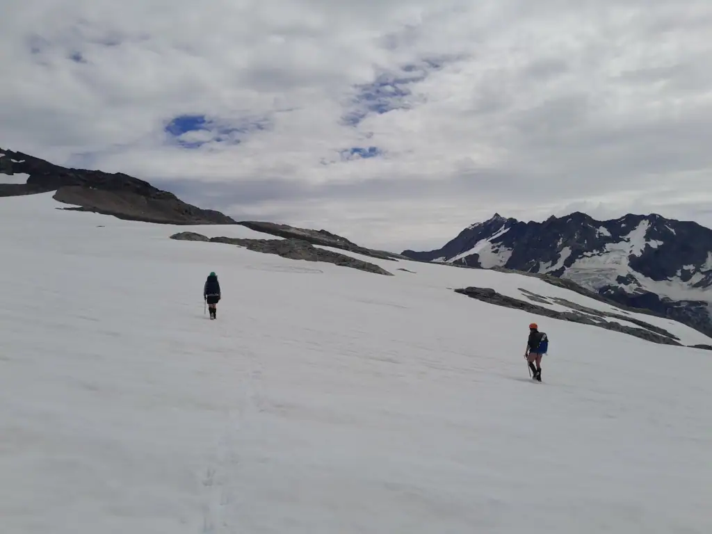 Two alpine hikers with backpacks wearing helmets and carrying ice-axes navigate across a vast, snow-covered plain during the Annette Plateau traverse, Aoraki/Mt Cook National Park