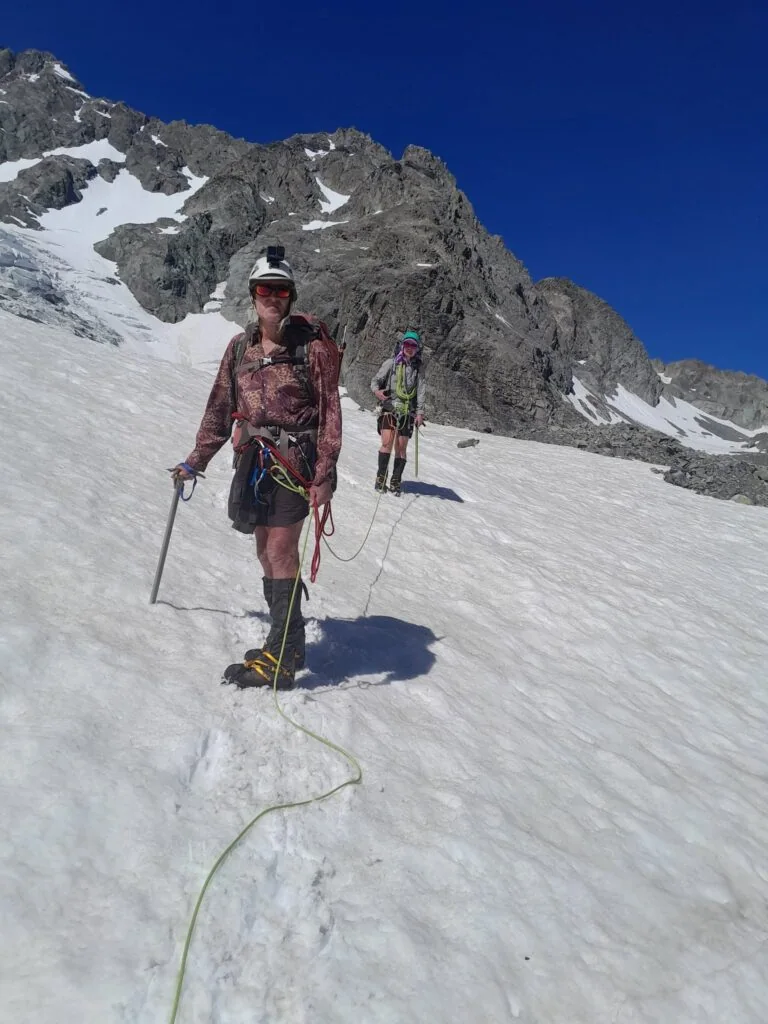 Two mountaineers roped up for safety while descending a steep snowfield on the Ashburton Glacier, equipped with helmets, ice axes, and climbing harnesses against a vivid blue sky in the Arrowsmith Range, Hakatere Conservation Park, Canterbury