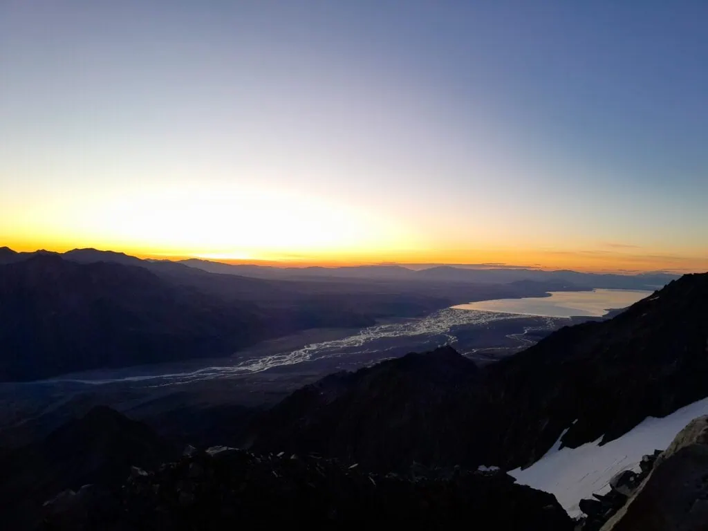 A wide scenic landscape at dawn from Mt Annette on the Annette Plateau showing the bright yellow horizon reflecting onto the winding channels of the braided Tasman River valley flowing into Lake Pukaki, Aoraki/Mt Cook National Park