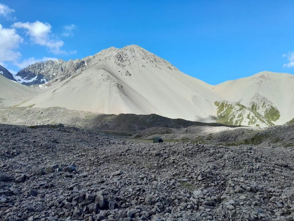 The small green Cameron Hut nestled in a massive, rocky glacial basin of the Cameron Glacier beneath the steep scree slopes of the Arrowsmith Range, including The Marquee, Hakatere Conservation Park, Canterbury