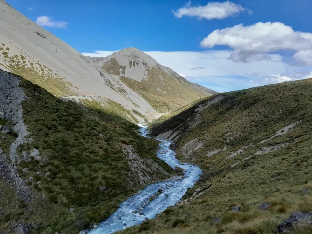 A high-angle view from a steep river terrace looking down a steep valley where the swollen blue Cameron River carves through massive scree mountain slopes. Located on the Cameron Hut Route in Hakatere Conservation Park, Canterbury