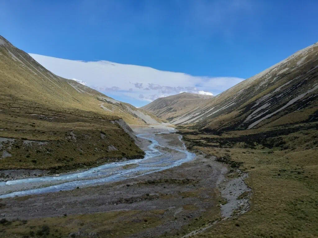 The braided Cameron River flowing through a vast, deep alpine valley with golden tussock and scree-covered mountains under a striking blue and white sky. Located on the Cameron hut Route, Hakatere Conservation Park, Canterbury