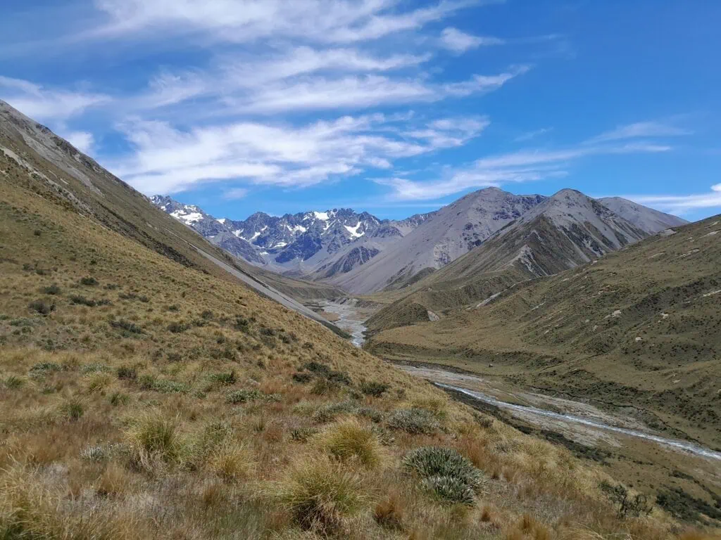 A sunny afternoon view looking down the golden tussock slopes of the Cameron Valley, with a winding river below and the jagged, snow-capped Arrowsmith Range in the distance. Historic Musterers' Hut, Highland Hut, is visible on the far bank of the Cameron River. Located in Hakatere Conservation Park, Canterbury