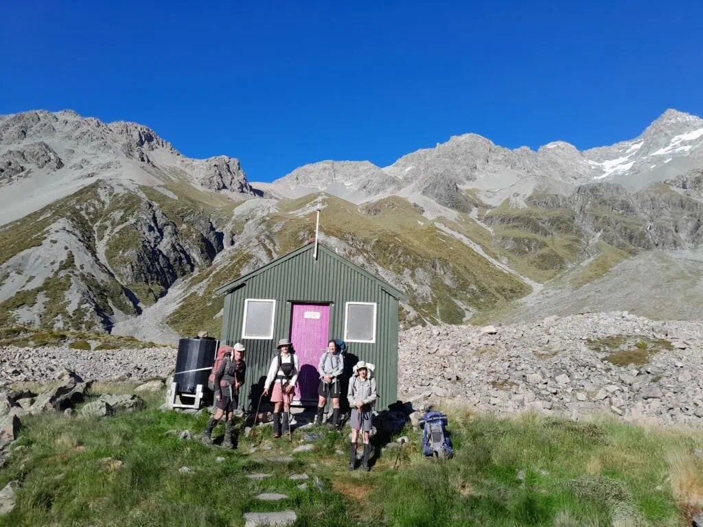 Four trampers with backpacks standing outside the green, purple-doored Cameron Hut at the head of the Cameron Valley, nestled below the towering scree slopes and rugged peaks of the Arrowsmith Range, Hakatere Conservation Park, Canterbury