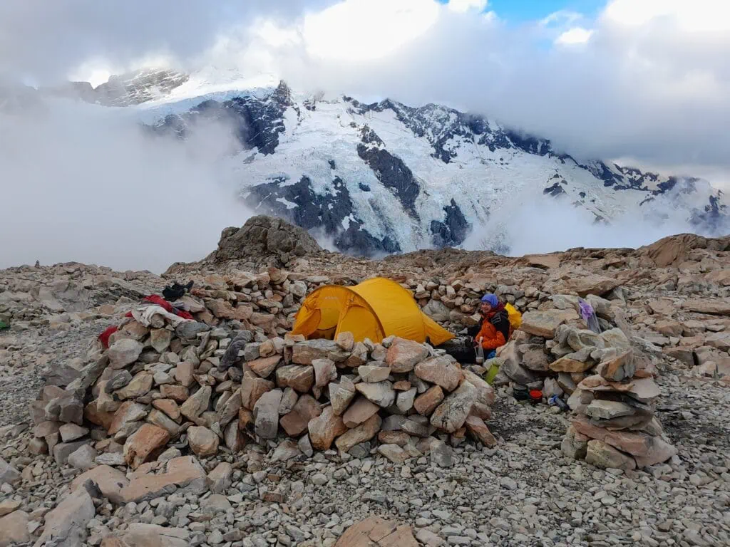 An alpine tramper sitting next to a yellow tent inside a rock wall wind site at Mueller Hut, with low-lying clouds swirling around the hanging glaciers behind them & Mt Sefton, Aoraki/Mt Cook National Park