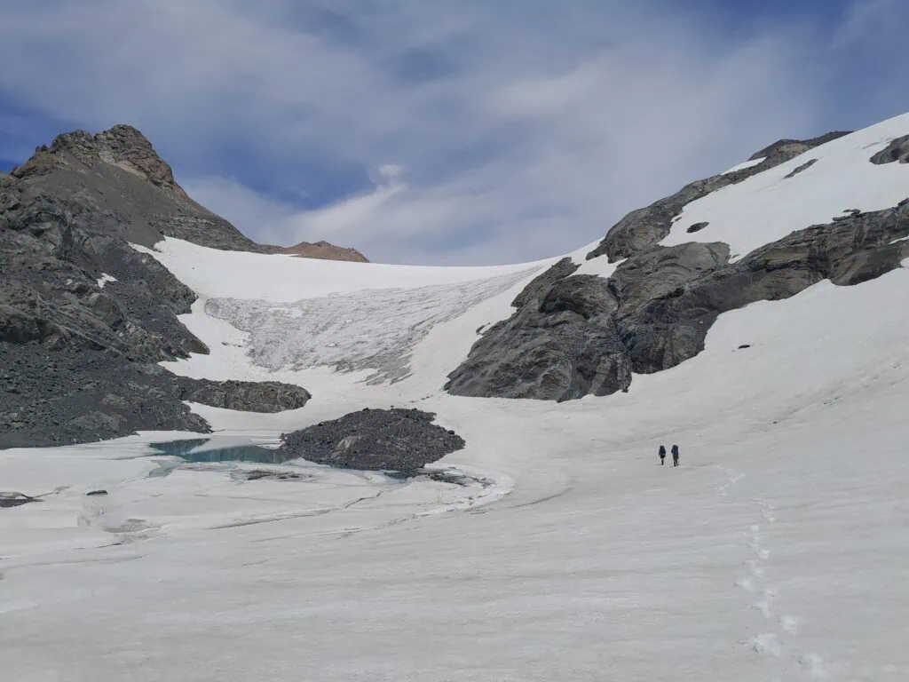 Two mountaineers with large backpacks ascending a steep snow slope towards the Annette Plateau under a partly cloudy sky, Aoraki/Mt Cook National Park