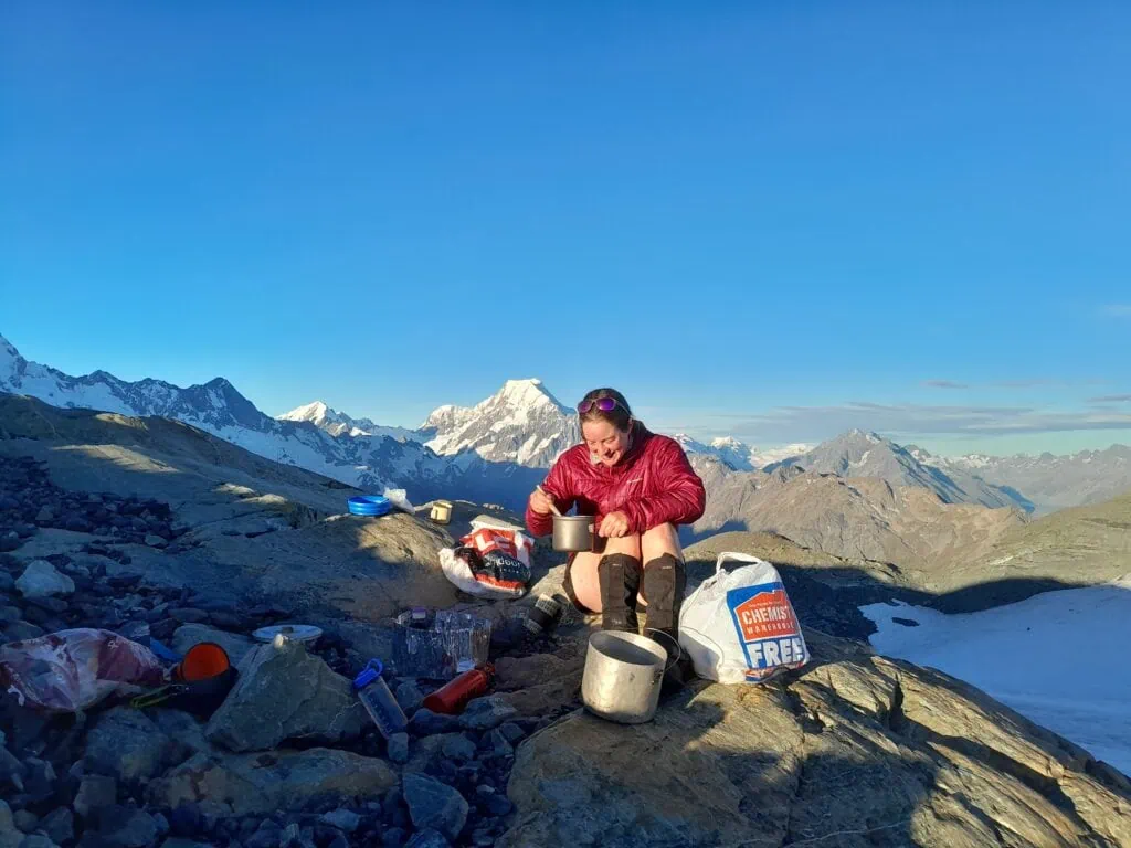 A hiker sits on a rocky ridge cooking a meal on a camping stove, with a stunning backdrop of Aoraki/Mount Cook and a clear blue sky on the Annette Plateau in Aoraki/Mt Cook National Park