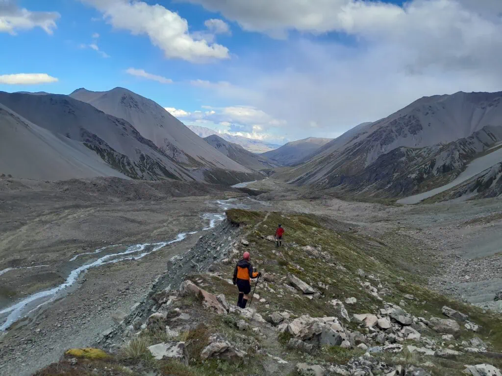 Two hikers descending from Cameron Glacier a rocky, tussock-covered ridge route back into the wide glacial basin of Cameron Hut in the Arrowsmith Range in Hakatere Conservation Park, Canterbury.