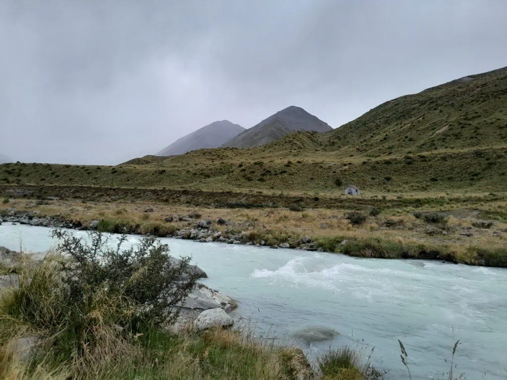 The fast-flowing, milky-blue waters of a swollen Cameron River under overcast skies, with a small historic musterers’ hut, Highland Hut, visible on the far river flats. Located in Hakatere Conservation Park, Canterbury