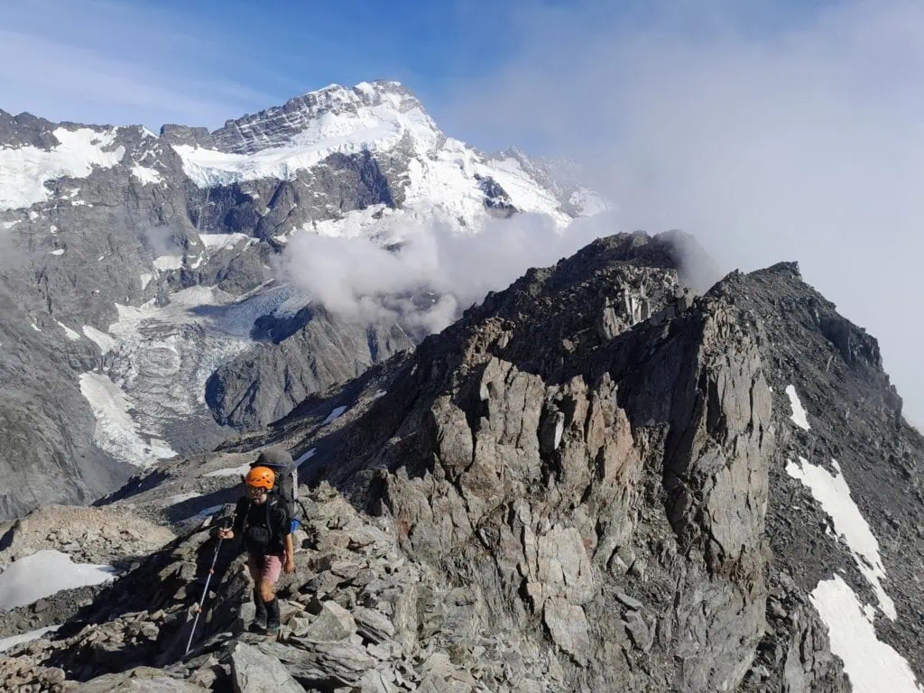 A mountaineer wearing an orange helmet and backpack traverses a rugged, rocky ridge line towards Mt Kitchener & Annette Plateau with a massive, snow-covered Mt Sefton in the background, Aoraki/Mt Cook National Park