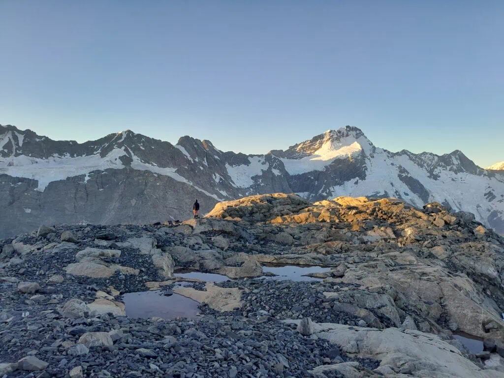 A lone tramper on the Annette Plateau standing amongst rocky alpine tarns on a ridge, looking out at the snow-capped peak of Mt Sefton catching the early evening sun, Aoraki/Mt Cook National Park.