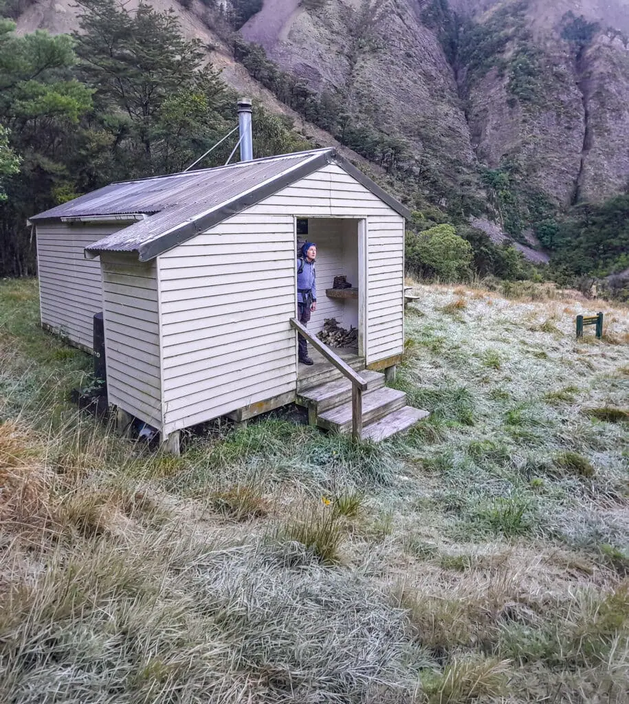 A tramper with a backpack standing in the doorway of the white weatherboard Isolation Hut on a frosty morning, in Isolated Hill Reserve in the Seaward Kaikōura Range
