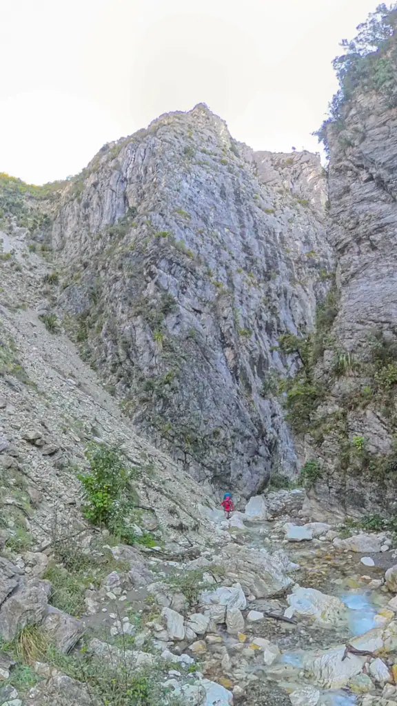 A tramper walking up a shallow stream bed in Waimā River framed by massive, sheer limestone cliffs leading into Sawcut Gorge, Isolated hill Scenic Reserve in Seaward Kaikōura Range.
