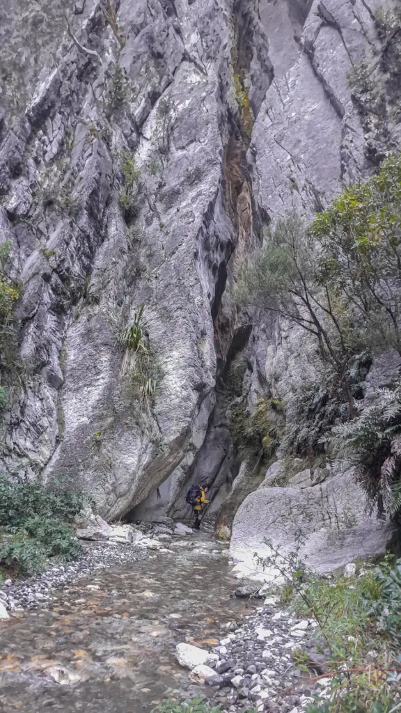 A tramper in a yellow rain jacket standing in the rocky creek bed at the deep limestone exit of Sawcut Gorge,in Isolated Hill Reserve in the Seaward Kaikōura Range