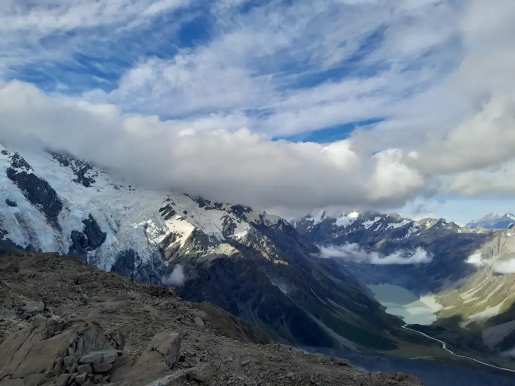 High-angle panoramic view looking down into the valley at Hooker Lake and its outflow river, surrounded by steep glacial mountain faces in Aoraki/Mt Cook National Park.