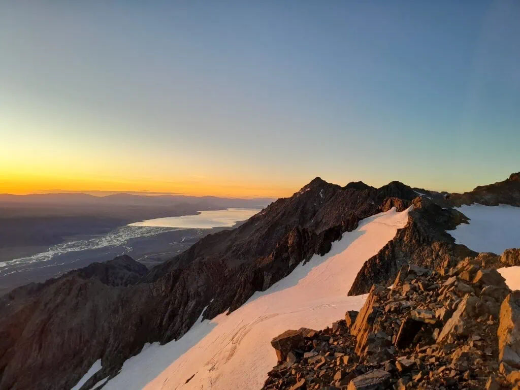 An elevated sunrise perspective from Mt Annette on the Annette Plateau showing the early morning sun illuminating a rocky, snow-lined ridge overlooking the long expanse of Lake Pukaki and the braided Tasman River, Aoraki/Mt Cook National Park