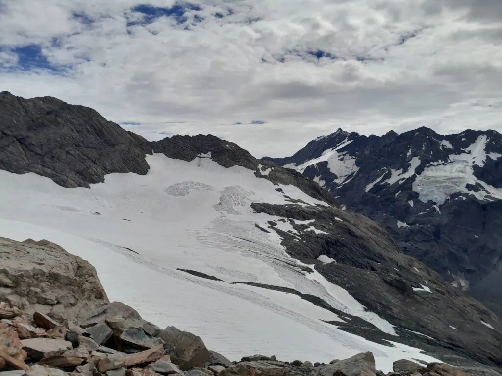 A wide view of the snow-packed Metelille Glacier with crevasses nestled between dark, rugged rock faces under a cloudy sky. Mueller Glacier lies far below it. Located in Aoraki/Mt Cook National Park