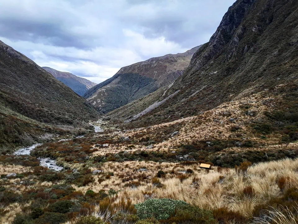 The tiny orange and black timber Minchin Biv nestled in a vast alpine valley of golden tussock and low scrub under a cloudy sky above Poulter Valley, Arthur's Pass, Canterbury
