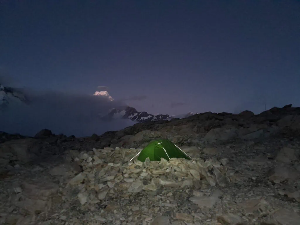 A green tent pitched within a protective rock wall under a dark twilight sky, with the snow-covered peak of Aoraki Mt Cook visible in the distance, Aoraki-Mt Cook National Park