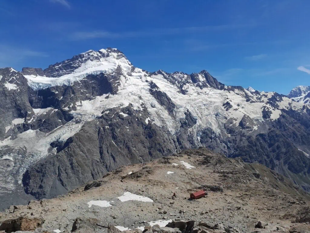 A high-angle view from Mt Ollivier looking down at the iconic red Mueller Hut situated on a rocky plateau, with a massive snow-covered Mt Sefton looming behind it under a blue sky, Aoraki/Mt Cook National Park