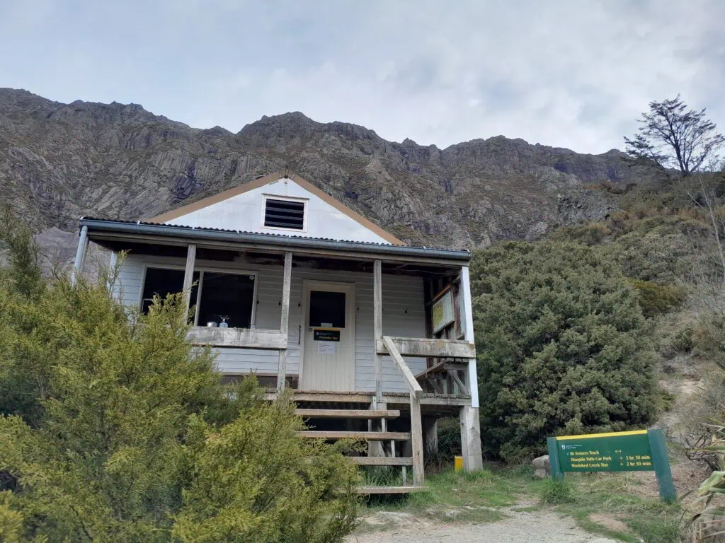 The wooden DOC Pinnacles Hut surrounded by native bush with a massive rocky mountain face towering in the background, Mt Somers Area, Hakatere Conservation Park, Canterbury