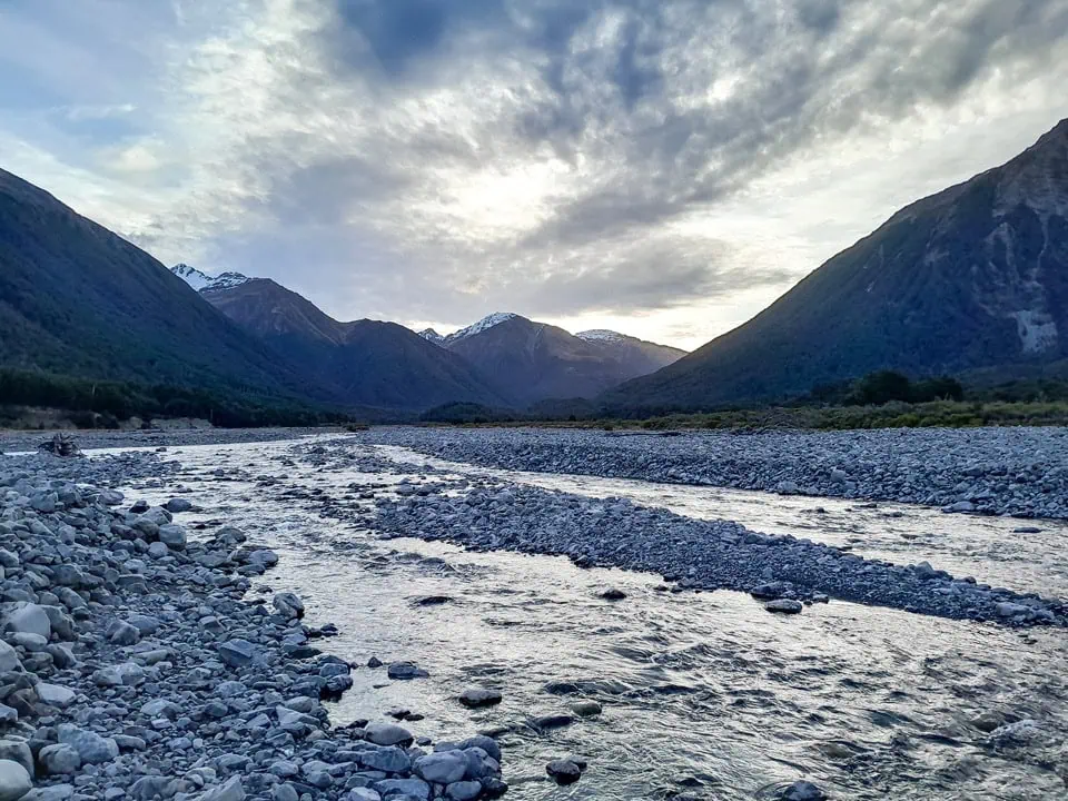 A wide, braided, gravelly riverbed of the Poulter River with flowing water channels snaking toward forested hills and mountain valleys at sunset near Poulter Hut in Arthur's Pass, Canterbury