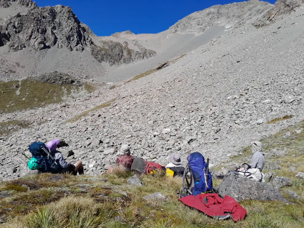 A group of trampers sitting with their backpacks on a tussock and scree slope, taking a rest break while looking up at a massive scree basin above Cameron Hut in the Arrowsmith Range while on a trans-alpine tramp to Ashburton Glacier, Hakatere Conservation Park, Canterbury.