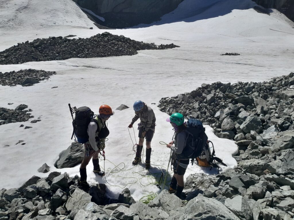 A team of three mountaineers in helmets and backpacks attaching a climbing rope together on the ice of the Ashburton Glacier, preparing for a crevasse crossing. Located in Hakatere Conservation Park, Canterbury