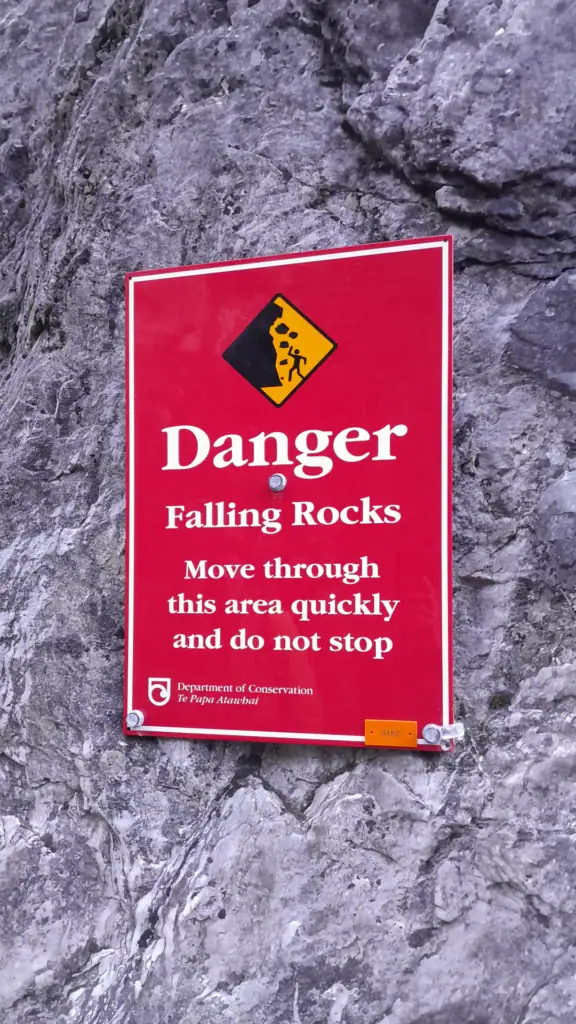 A close-up of a red Department of Conservation (DOC) warning sign for falling rocks mounted on a steep limestone cliff face in Sawcut Gorge, Isolated Hill Scenic Reserve, Seaward Kaikōura Range
