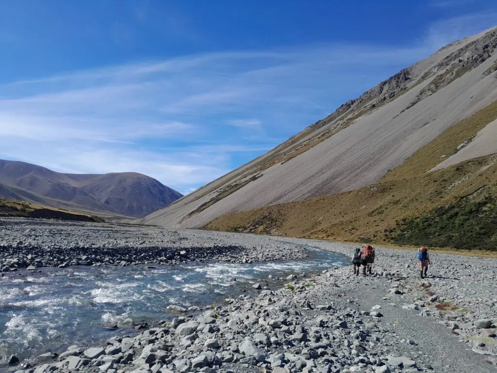 Four hikers with backpacks hiking along the rocky, gravelly banks of the fast-flowing South Branch Ashburton River/Hakatere beneath a towering scree mountain slope. Near Top Hut, Hakatere Conservation Park, Canterbury