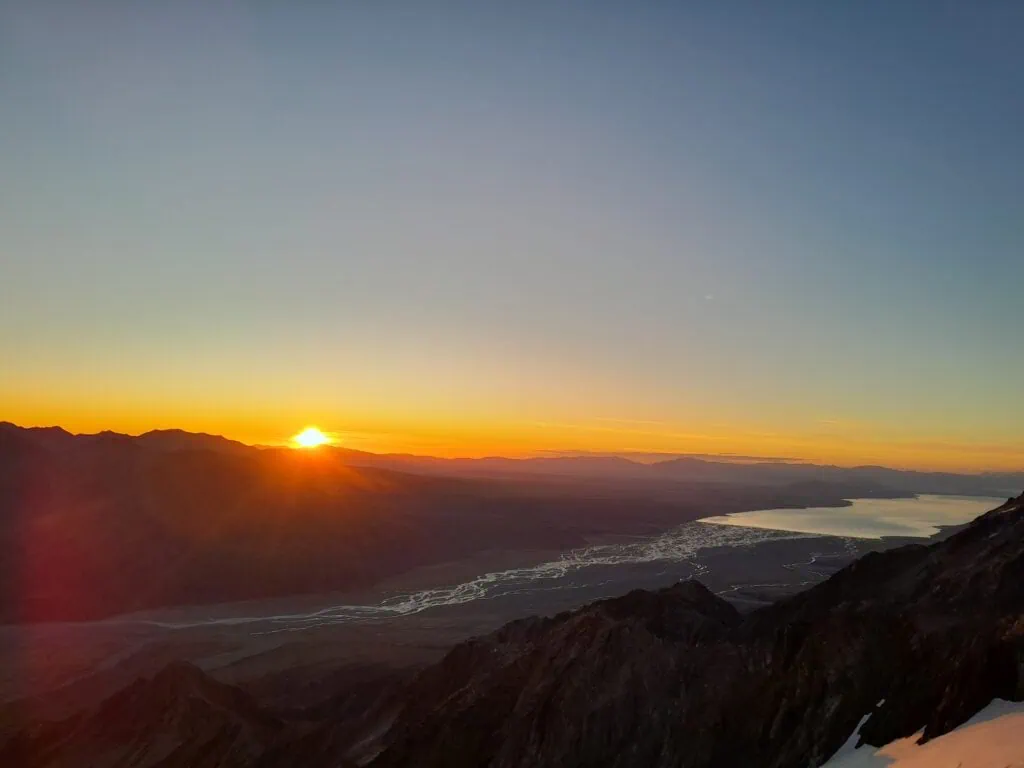 The sun bursts over the horizon of the Burnett Mountains, casting rays of light over the Tasman Valley containing the braided Tasman River and Lake Pukaki during an alpine sunrise from Mt Annette on the Annette Plateau in Aoraki/Mt Cook National Park.