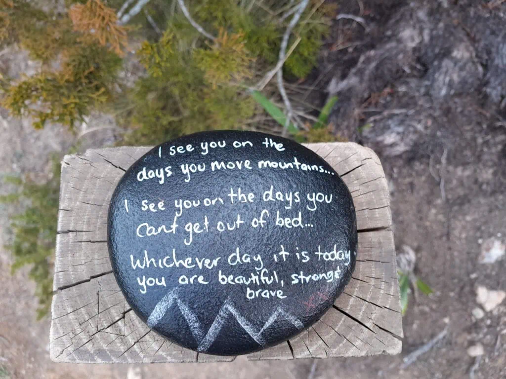 A smooth black inspirational rock with white handwritten text resting on a wooden post along a Mt Somers track in Hakatere Conservation Park, Canterbury.