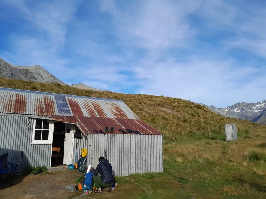 A hiker sorting gear outside the rustic, corrugated-iron Top Hut in the Hakatere Conservation Park on a clear, sunny morning, Canterbury