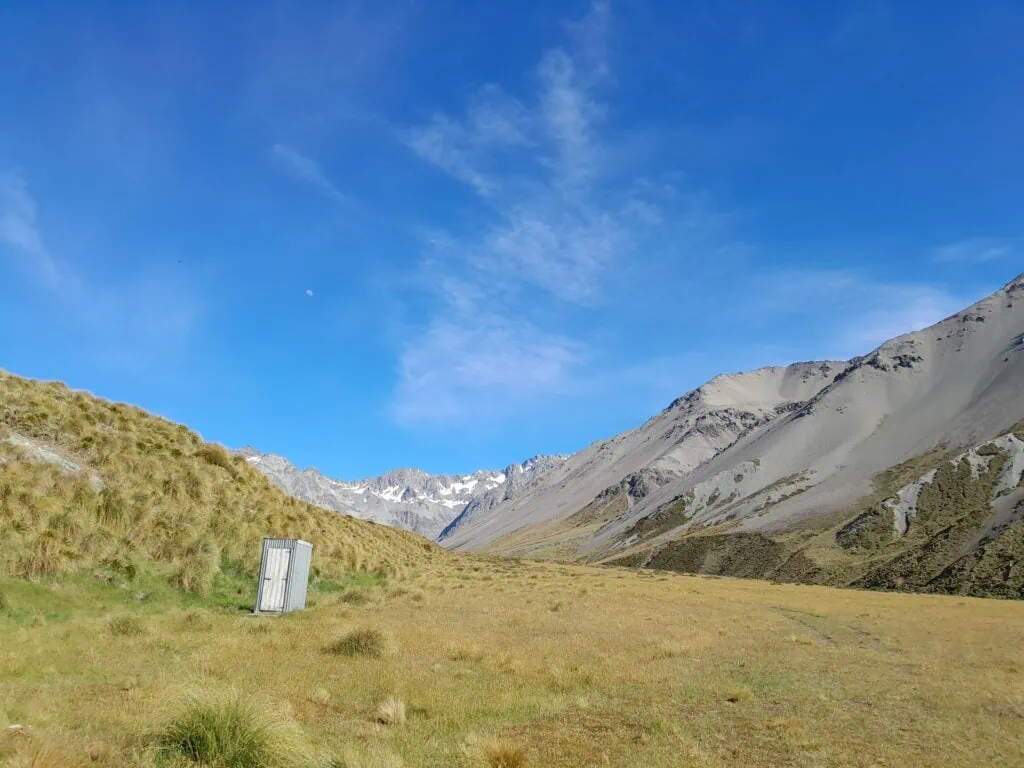 A lone corrugated-iron longdrop toilet standing in a vast, golden tussock valley with the dramatic scree slopes and snow-capped peaks of the Arrowsmith Range in Hakatere Conservation Park, Canterbury