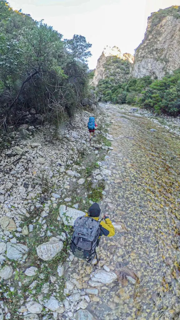 Two hikers, seen from an elevated angle, walking upstream along the shallow gravel bed of the scenic Waimā River to get to Sawcut Gorge in Isolated Hill Reserve in the Seaward Kaikōura Range