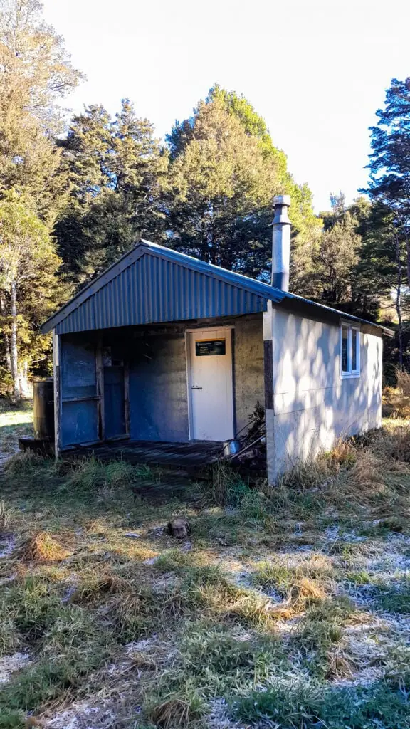 Zoo Hut, a simple corrugated iron and wooden backcountry DOC hut, sitting on a frosty grass clearing surrounded by native forest in Isolated Hill Reserve in the Seaward Kaikōura Range