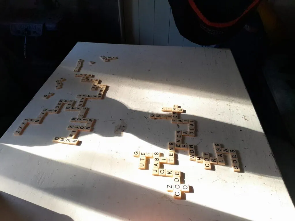 Bananagrams letter tiles spread across a white wooden table inside a Department of Conservation Hut, Zoo Hut, in Isolated Hill Reserve in the Seaward Kaikōura Range, with dramatic morning shadows.