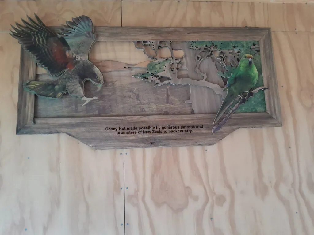 A decorative wooden artwork inside Casey Hut in the Poulter Valley featuring a carved kea and an orange-fronted kākāriki, dedicated to the patrons of the New Zealand backcountry, Arthur's Pass, Canterbury