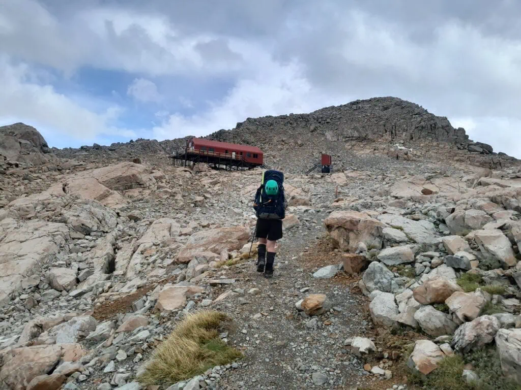 A tramper with a backpack & helmet walking up a rocky alpine trail toward the iconic red Mueller Hut perched on a mountain ridge, Aoraki-Mt Cook National Park