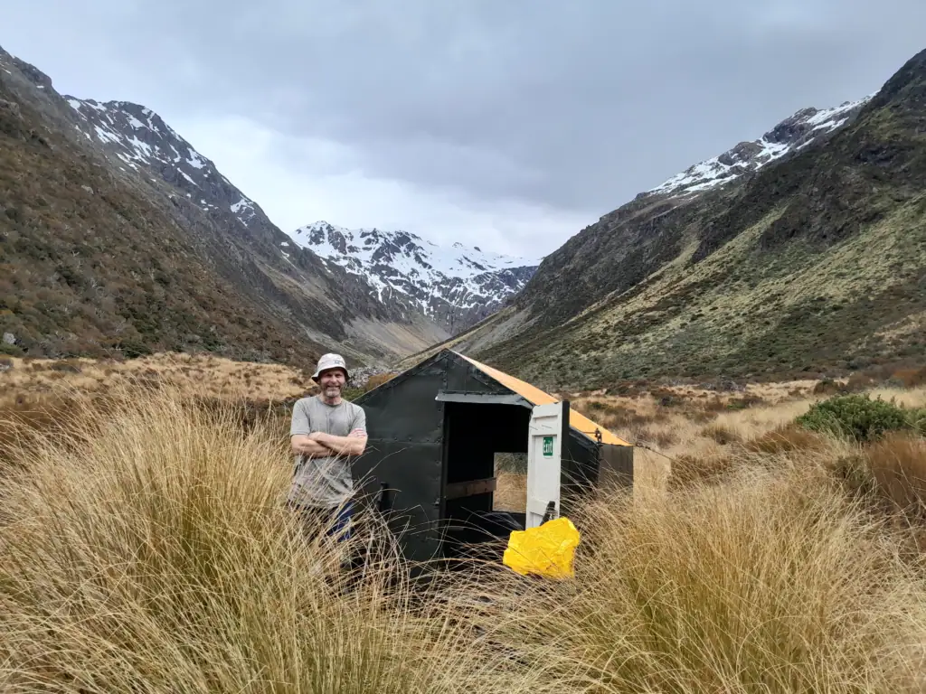 A smiling male tramper standing with arms crossed next to the small black and orange Minchin Biv surrounded by tall tussock grass and snowy mountain peaks, Arthur's Pass, Canterbury