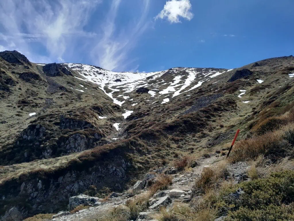 Looking up the rugged, steep Mt Somers Summit Track toward a snow-covered alpine ridge with a DOC orange trail marker pole, Hakatere Conservation Area, Canterbury