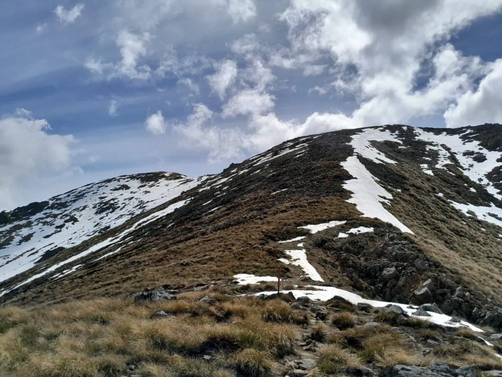 A steep, rocky ridge with patches of spring snow along the Mt Somers Summit Track under a cloudy blue sky, Hakatere Conservation Area, Canterbury