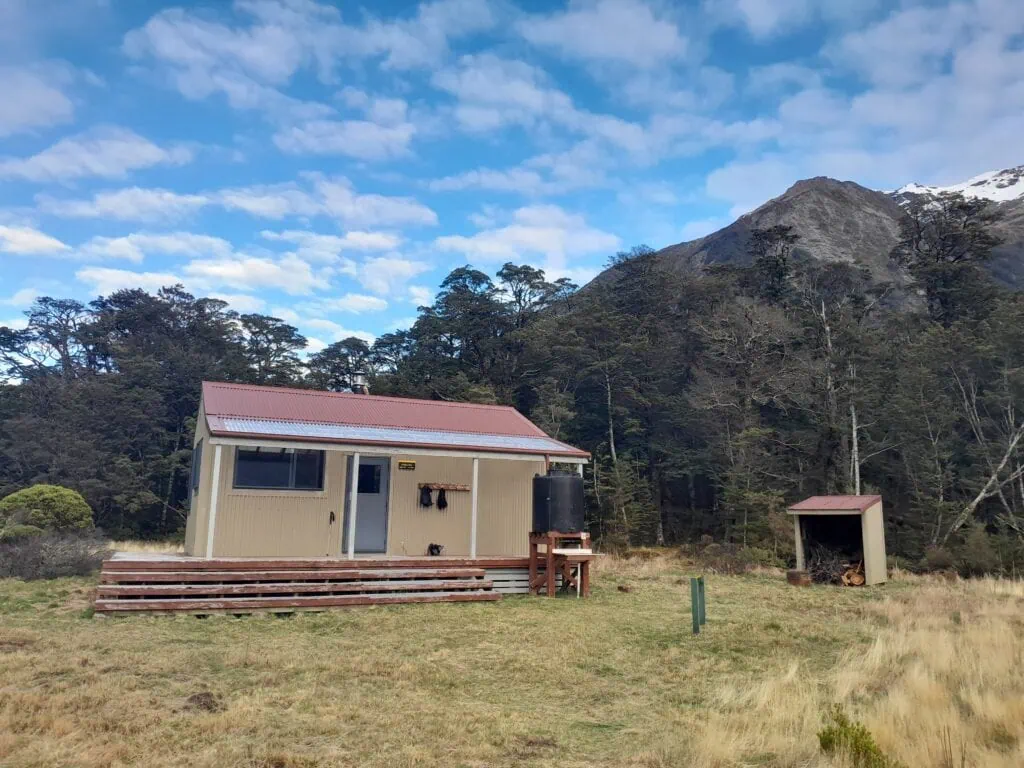 Poulter Hut with a red-brown tin roof sitting on a grassy flat in front of a dense beech forest and mountains under a blue sky, Arthur's Pass National Park. On the way to Lake Minchin