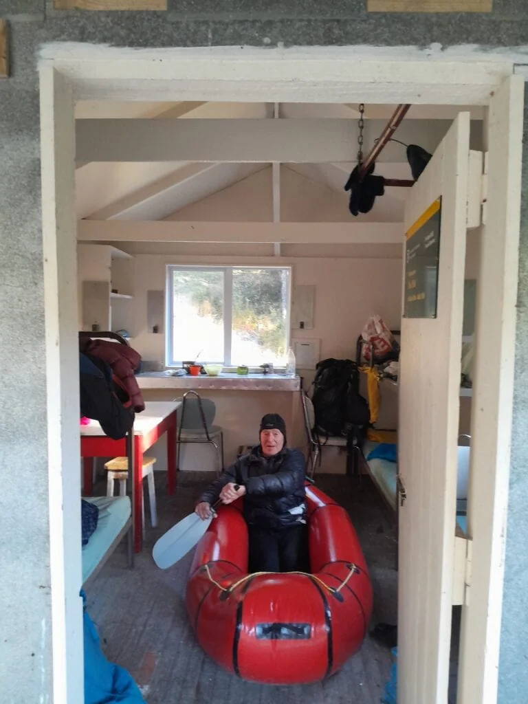 A tramper sitting in an inflated red packraft with a paddle inside Zoo Hut, preparing for the Waimā Gorge. Located in Isolated Hill Reserve in the Seaward Kaikōura Range