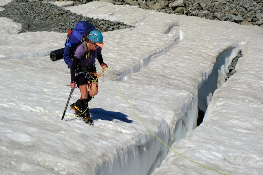 A female mountaineer with a large backpack and helmet carefully holding a rope while stepping over a deep, open crevasse on the Ashburton Glacier, Arrowsmith Range, Hakatere Conservation Park, Canterbury