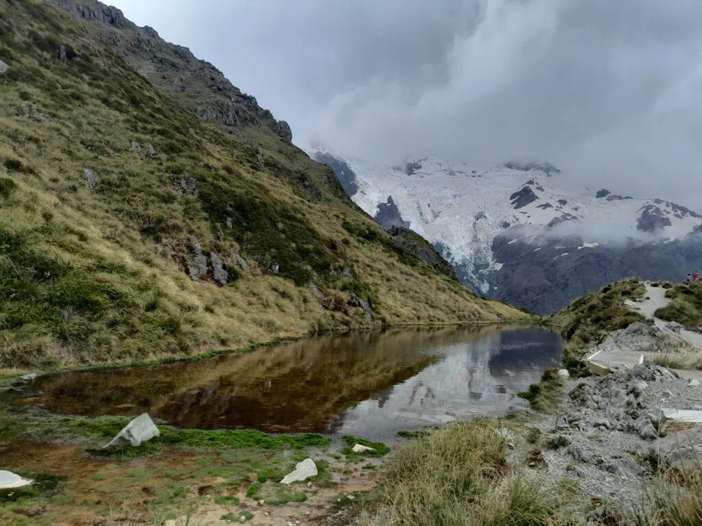 The calm water of Sealy Tarns reflecting the surrounding green hillsides and clouds, with a massive glacier-covered mountain face looming in the background, Mt Sefton. Located in Aoraki/Mt Cook National park on the climb to Mueller Hut & Annette Plateau