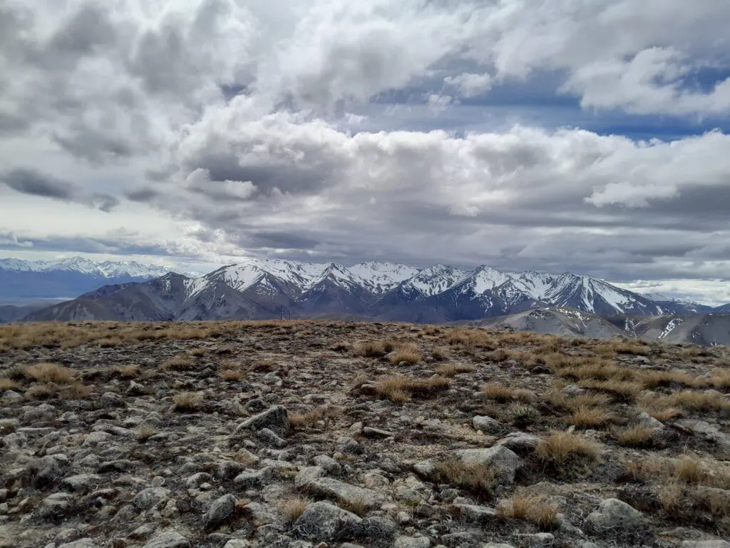 Wide summit views from Mount Somers showcasing the rocky alpine plateau and a massive backdrop of snow-capped mountains of Hakatere Conservation Park, Canterbury