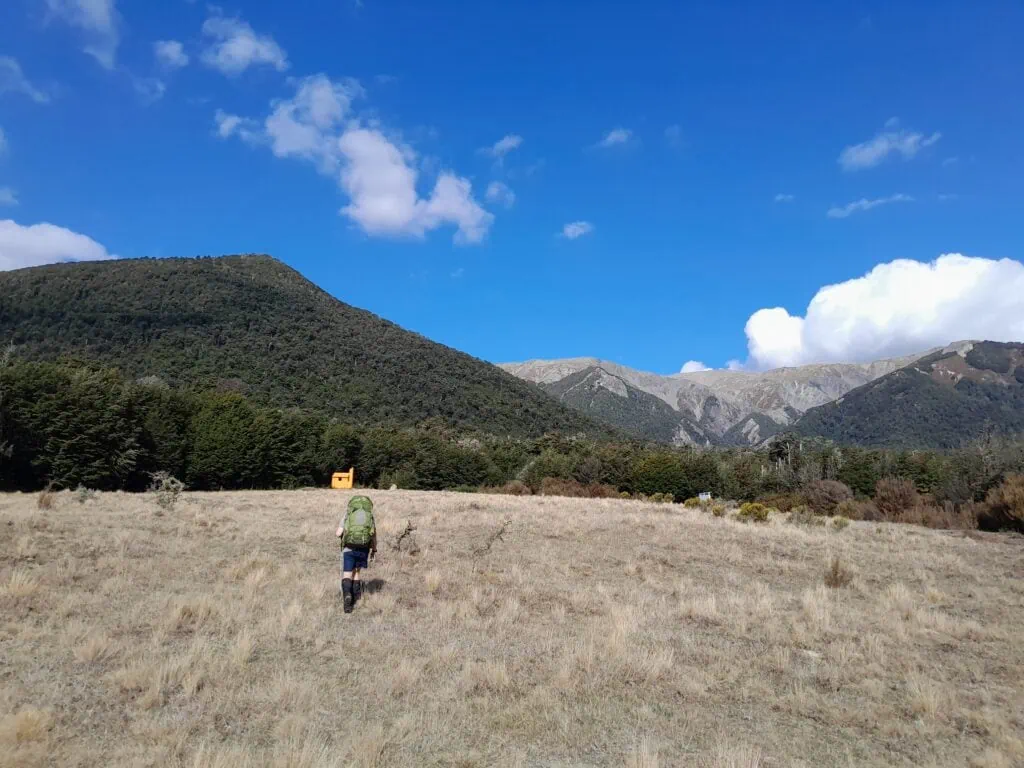 A tramper carrying a large green backpack walks up a wide, dry grassy river terrace toward a tiny orange structure in the distance that is Turnbull Biv in the Poulter Valley, Arthur's Pass.